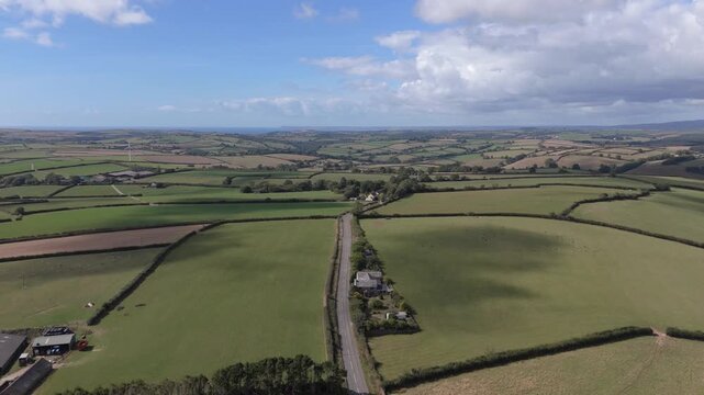 A bright, sunny day with scattered clouds over the rolling green fields and hedgerows of the Cornish countryside near Looe. The camera rises, revealing a patchwork of agricultural land.