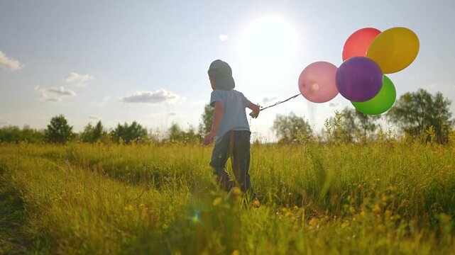 Boy runs through meadow holding colorful balloons at sunset. Child plays with balloon bunch in field outdoors. Happy boy with balloons runs in meadow. Kid enjoys balloon fun in nature field.