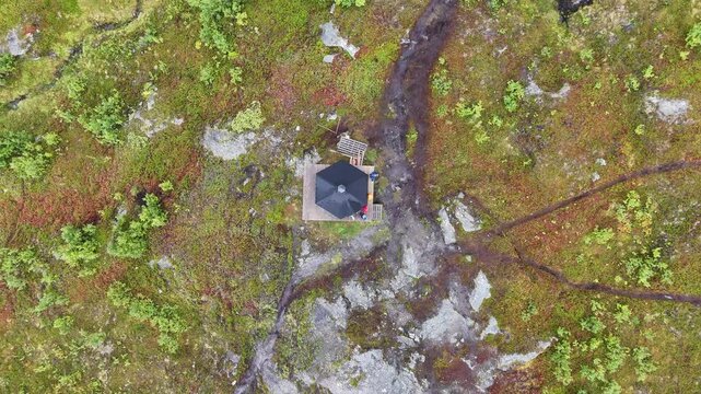 Aerial view of a hexagonal structure on a wooden deck surrounded by rocky mossy terrain with people standing nearby in Lyngseidet, Troms, Norway.