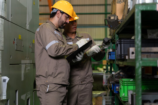 Industrial worker in a hard hat and protective clothing checks a clipboard beside factory machinery, representing equipment inspection, manufacturing operations, technical work, and workplace safety.