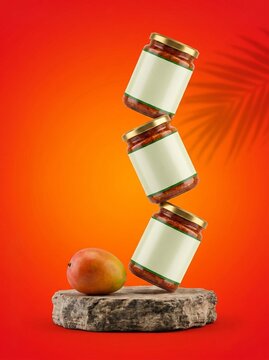 Three jars of mango pickle stacked on a stone pedestal with a fresh mango against a vibrant orange and red background.