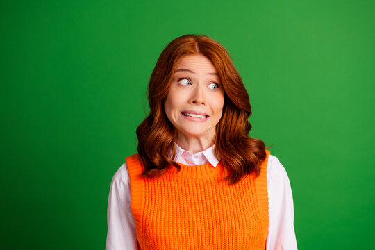 Nervous redhead woman in orange sweater vest with anxious expression against a green background