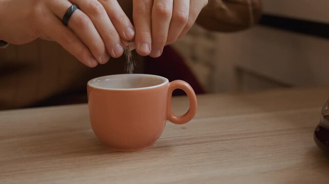 Close up of person hands tearing open and pouring sugar into orange ceramic cup on wooden table. scene features soft lighting with another filled mug visible in background emphasizing calm