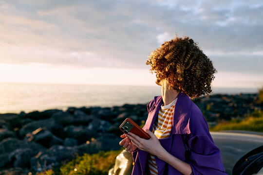 A young woman with curly hair stands beside a rocky coast at sunset, holding a phone and gazing into the distance, conveying a reflective mood and quiet curiosity, wearing a purple jacket