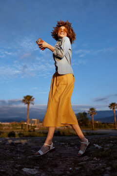 Joyful woman outdoors walking on a sunlit path, casual outfit and breeze in her hair, smile and relaxed movement against a clear blue sky and palm trees background