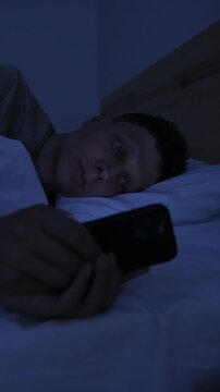 Man compulsively scrolling smartphone in bed at night, struggling with digital addiction, doomscrolling and inability to stop screen time, vertical
