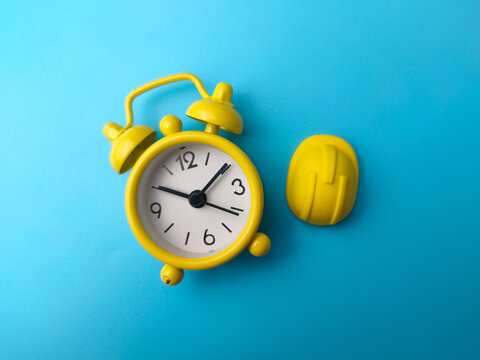 A mini yellow hard hat toy sits beside a yellow alarm clock on a blue background