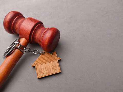A wooden house keychain hangs from the head of a wooden judge gavel on a gray surface