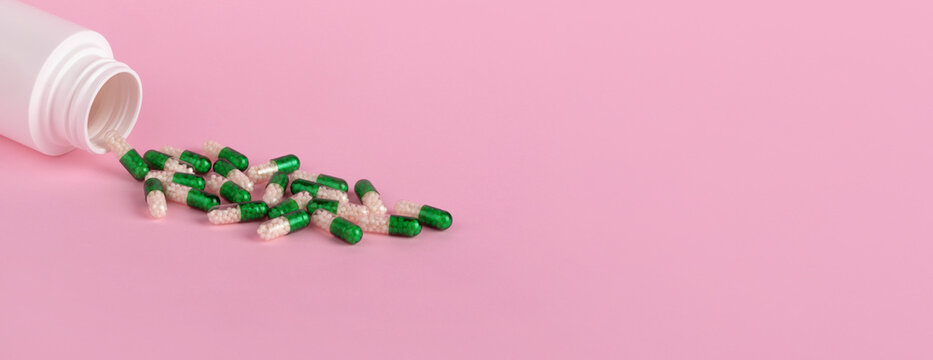 Open white bottle with green and white capsules spilling on pastel pink surface, ideal for healthcare, pharmacy, and wellness concepts.