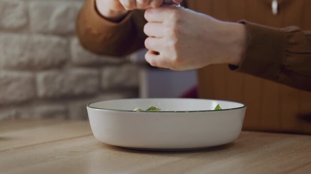 Close up of person seasoning food in white bowl on wooden table. Hands holding pepper grinder sprinkle salt over green leaves with stone wall and kitchen background visible