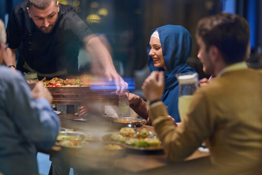 Smiling Woman in Hijab Enjoying Group Dinner at Restaurant While Server Presents Shared Platter