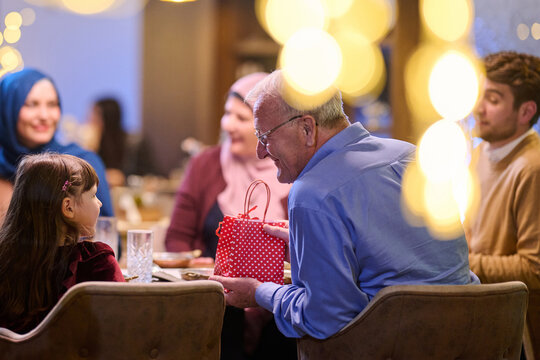 Grandfather Giving Gift To Granddaughter At Multigenerational Family Holiday Dinner With Festive Lights