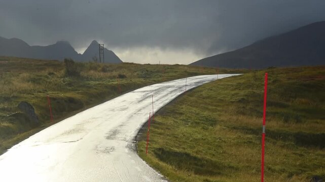 Wet mountain road marked with snow poles near Napp under a stormy sky, Lofoten, Norway