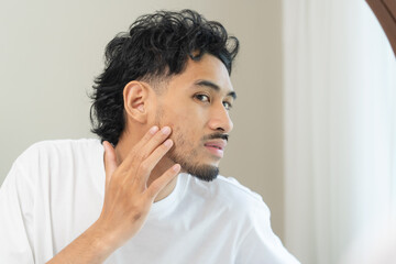 Handsome young Asian man looking in the mirror and checking his skin during morning routine at home