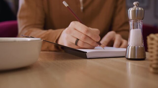 Close up of woman writing in notebook with soft lighting and calm expression. scene includes objects like salt shaker bowl on wooden table indicating indoor setting. Background is blurred