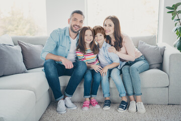 Fototapeta na wymiar Smiling family of four sitting together on a sofa in a bright living room, parents hugging two children for a cozy at-home portrait