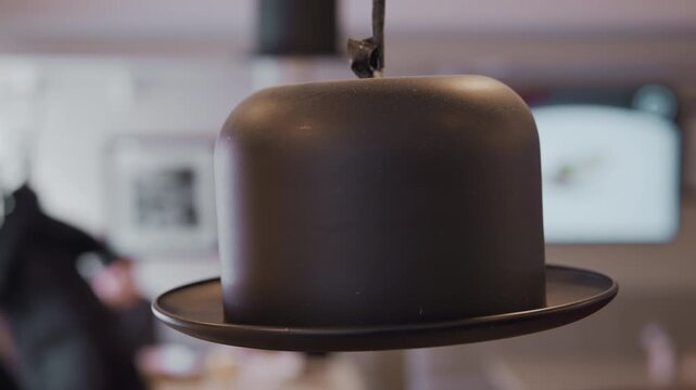 Close up of black bowler hat suspended from ceiling on round plate with soft lighting and shallow depth of field. background shows indoor setting with blurred people dining at tables framed