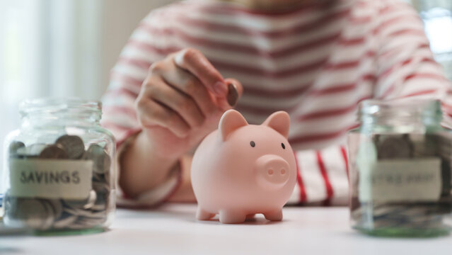 Woman placing a coin into a piggy bank beside savings jars, symbolizing personal finance, budgeting, emergency funds, and long-term wealth planning.