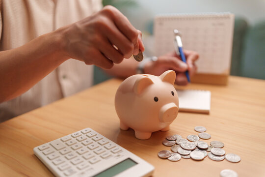 Asian man puts a coin into a piggy bank beside a calculator and notes at home, representing savings goals, budgeting