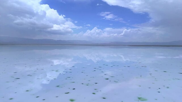 Slow forward‑moving aerial drone footage over Chaka Salt Lake in Qinghai, mirror‑like salt flat reflecting fluffy clouds, blue sky and distant hazy mountain range