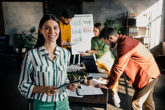 Young businesswoman leading a collaborative team meeting in a modern office while colleagues analyze charts, discuss data and work on laptops during a creative planning session