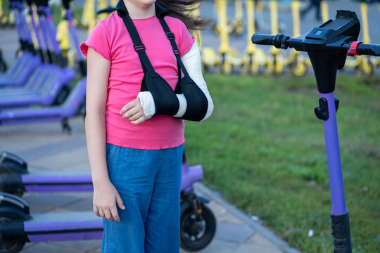 Unrecognizable girl with broken arm in plaster cast and sling standing near parking lot of electric scooters, child injury and urban safety concept