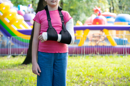 Girl with bone fracture in full arm cast and sling standing near inflatable trampoline. Childhood injury and safety concept