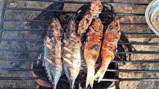 Fresh fish being grilled on a traditional charcoal stove with smoke rising, outdoor cooking process.