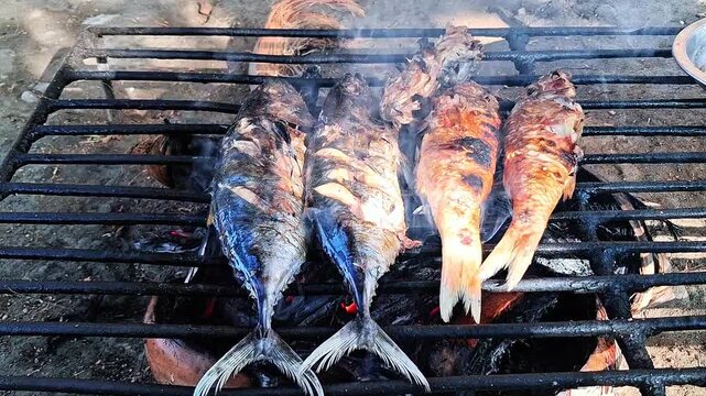 Fresh fish being grilled on a traditional charcoal stove with smoke rising, outdoor cooking process.