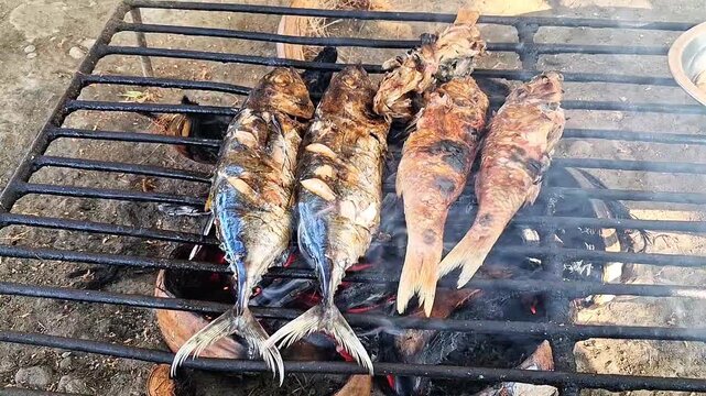 Fresh fish being grilled on a traditional charcoal stove with smoke rising, outdoor cooking process.