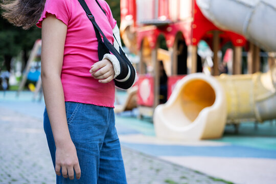 Girl with bone fracture in full arm cast and sling standing near playground summer. Childhood injury and safety concept