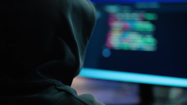 Anonymous hacker in a dark hoodie typing on a computer in a dimly lit room. Blurred colorful programming code on the monitor suggests cybercrime, cybersecurity breaches, or software development.