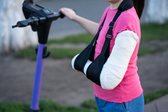 Unrecognizable girl with broken arm in plaster cast and sling standing near electric scooter, child injury and urban safety concept