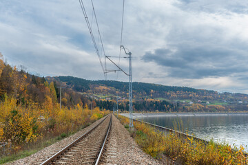 Electrified railway runs beside Jezioro Mucharskie near Stryszow, surrounded by colorful autumn forest and Beskid hills. Scenic rail transport landscape with reservoir shoreline, catenary system and m © Volha