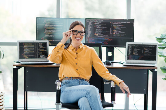 Smiling female software developer sitting at desk with multiple monitors and laptops coding in a bright modern home office
