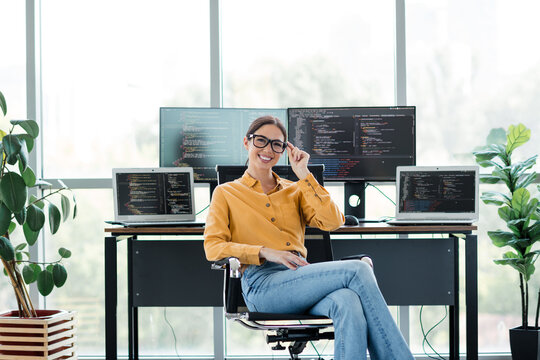 Female software developer smiling at desk with multiple monitors and laptops displaying code in bright modern home office workspace with plants