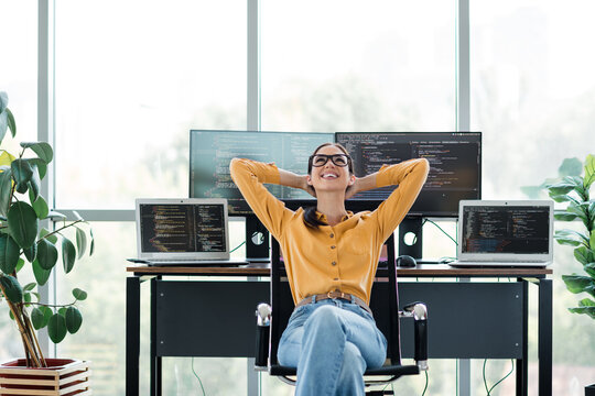 Relaxed female programmer leaning back at desk with multiple monitors and laptops displaying code in a bright modern home office workspace, enjoying a satisfied break