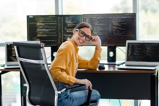 Female software developer smiling at a modern multi-monitor workstation while coding on laptop and desktop screens in a bright office