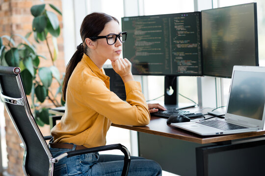 Female software developer concentrating on code at dual monitors in modern office workspace with laptop and ergonomic chair