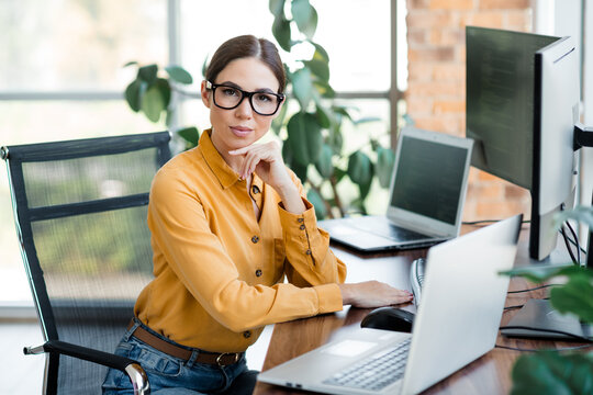 Professional businesswoman working at a computer in a bright modern office, wearing glasses and a mustard blouse while using laptop and desktop monitors