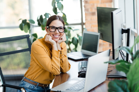 Smiling businesswoman working at laptop in modern bright office with multiple monitors and plants, relaxed professional wearing glasses and yellow blouse at desk