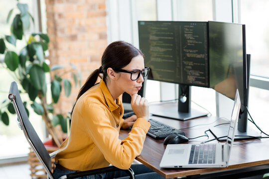 Focused woman programmer working at dual monitors and laptop in a modern office coding and debugging software during a concentrated development session