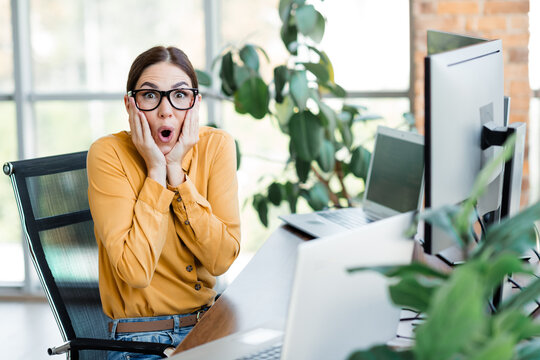 Surprised businesswoman at desk reacting to shocking news while working in modern office with laptop and monitor
