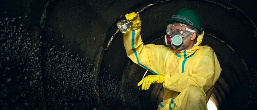 Hazmat Worker Examines a Collected Water Sample Inside an Industrial Drainage Tunnel While Wearing a Protective Suit, Respirator, Safety Helmet, Goggles, and Chemical-Resistant Gloves