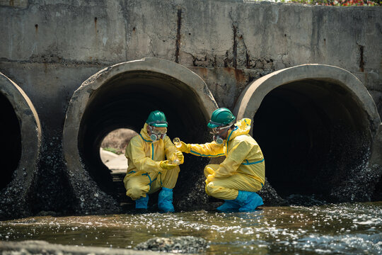 Environmental Workers Perform Wastewater Inspection Outdoors and Compare Contaminated Water Samples Beside Large Industrial Drainage Tunnels While Wearing Full Protective Equipment