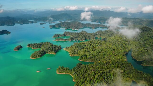 Aerial drone flight over KHAO SOK National Park in Thailand. Stunning views of Cheow Lan Lake's vibrant turquoise water surrounded by lush tropical jungle, iconic limestone karsts under morning clouds