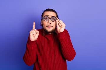 Naklejka premium Young handsome man with glasses wears a red sweater and poses playfully in a violet purple studio backdrop casual style fashion for advertising and shopping campaigns