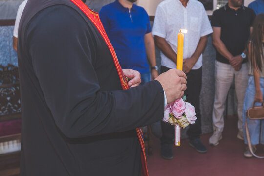 Orthodox priest holding burning candle and pink flowers during sacred religious ceremony with participants gathered around.