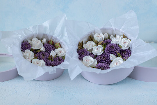 Floristry from Zephyr. Floral arrangement in round purple boxes featuring white roses and purple asters with decorative tissue paper on a light blue background