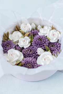 Floristry from Zephyr. Arrangement of white roses and purple chrysanthemums in a round box with tissue paper, set against a light blue background
